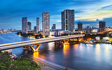 Thailand: Photo of a bridge over the Chao Phraya river in Bangkok, the bridge is illuminated and there are houses and skyscrapers in the background