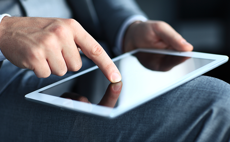Ein weißes Tablet liegt auf dem Oberschenkel eines Mannes in Anzug, er tippt darauf herum