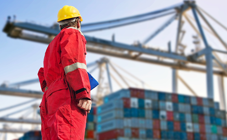 FCL: employee in red overall in port, with many containers and cargo cranes in the background