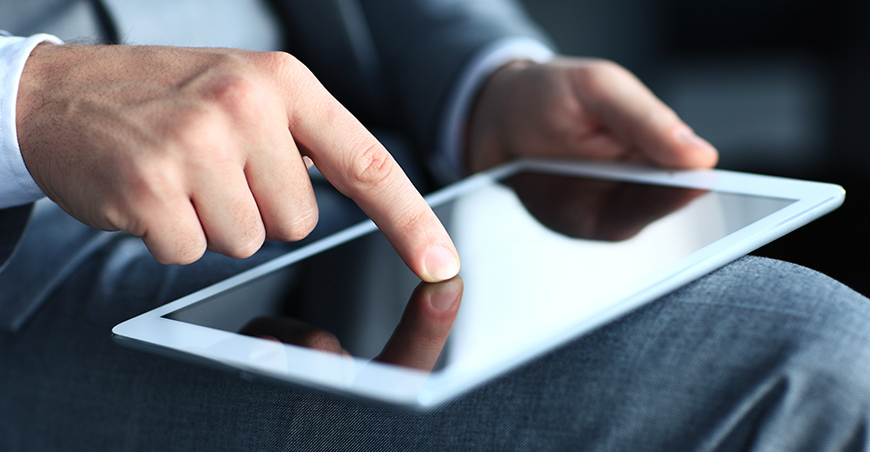 A white tablet lies on the thigh of a man in a suit, he is typing on it